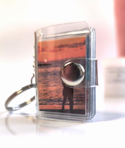 Mini-Fotobuch-Schlüsselanhänger mit einem Bild eines Paares bei einem Sonnenuntergang am Meer auf der Vorderseite, verschlossen mit einem silbernen Druckknopf.